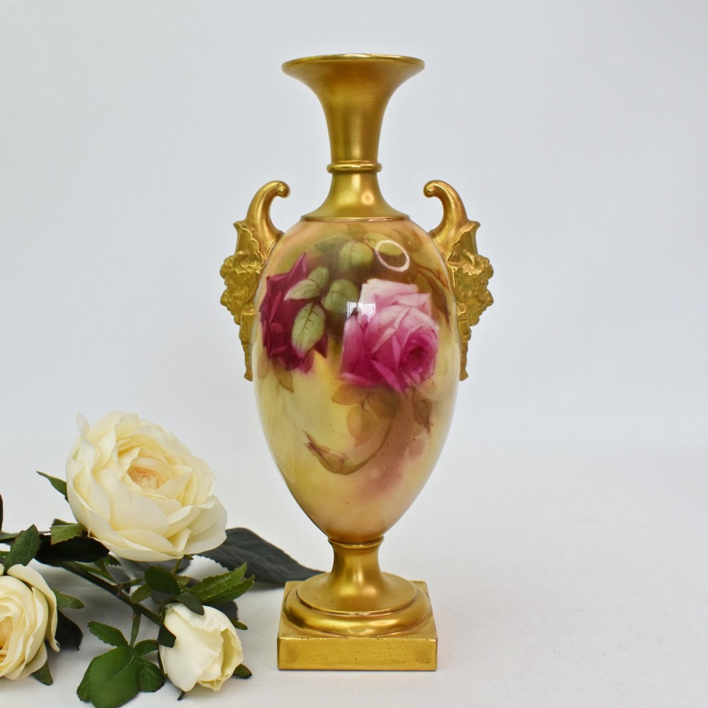 A hand-painted Royal Worcester porcelain vase decorated with pink and red roses, featuring gilded jester mask handles and a square base.