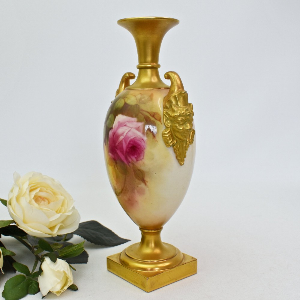 A hand-painted Royal Worcester porcelain vase decorated with pink and red roses, featuring gilded jester mask handles and a square base. View II