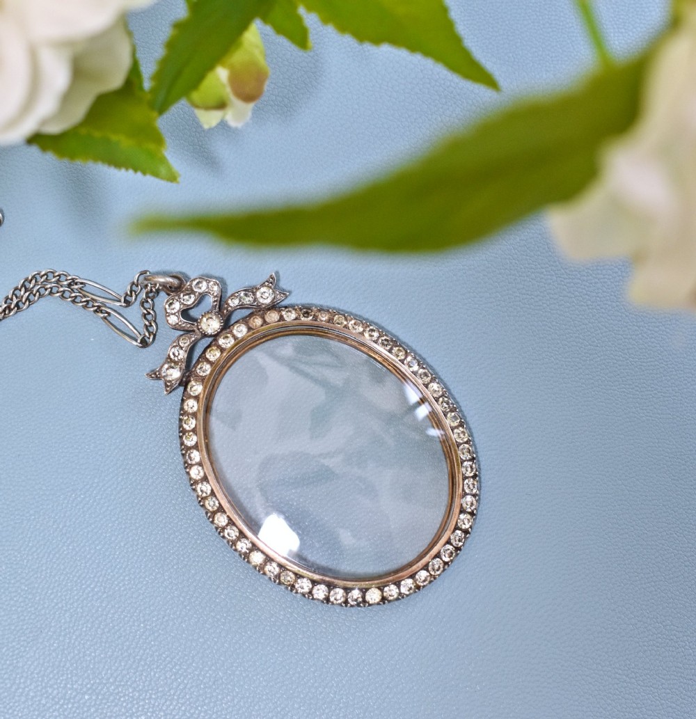 An oval glass portrait locket framed by a border of paste stones and topped with a silver ribbon bow motif on a chain.