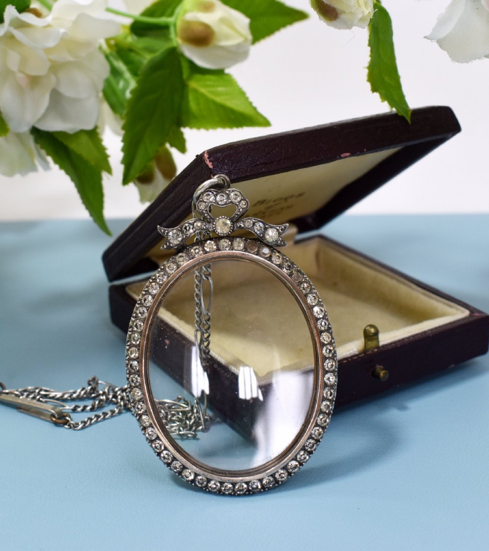 An oval glass portrait locket framed by a border of paste stones and topped with a silver ribbon bow motif on a chain. View II