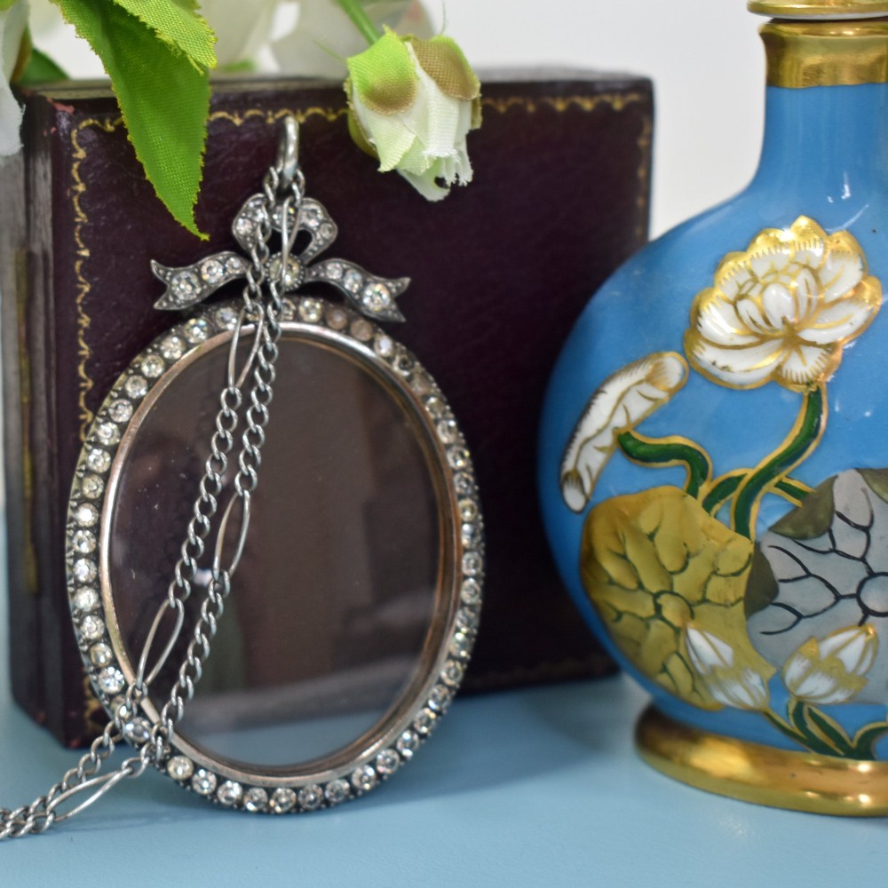 An oval glass portrait locket framed by a border of paste stones and topped with a silver ribbon bow motif on a chain. View III