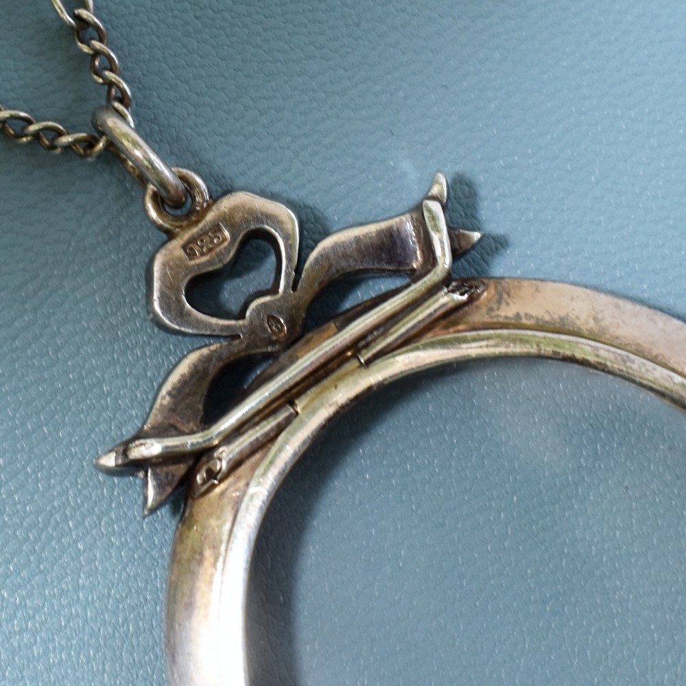 An oval glass portrait locket framed by a border of paste stones and topped with a silver ribbon bow motif on a chain. View IV