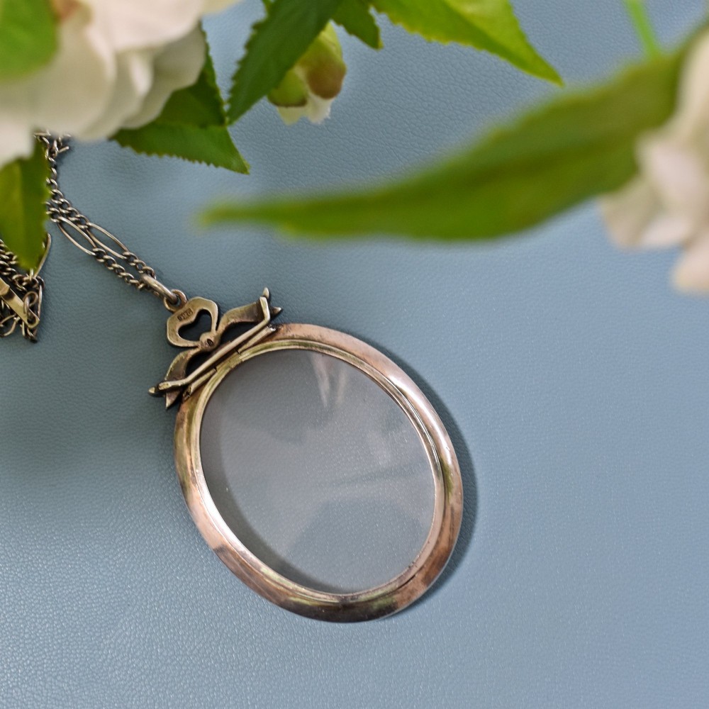 An oval glass portrait locket framed by a border of paste stones and topped with a silver ribbon bow motif on a chain. View V