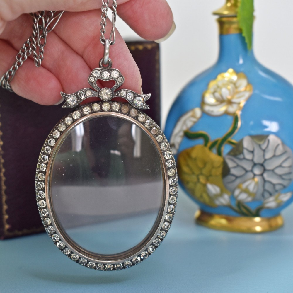 An oval glass portrait locket framed by a border of paste stones and topped with a silver ribbon bow motif on a chain. View VII