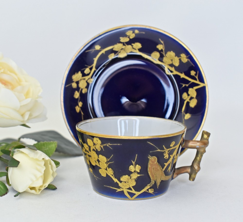 A dark cobalt blue porcelain coffee cup and matching saucer decorated with raised gilt floral branches, a perched bird, and a bamboo-style handle.