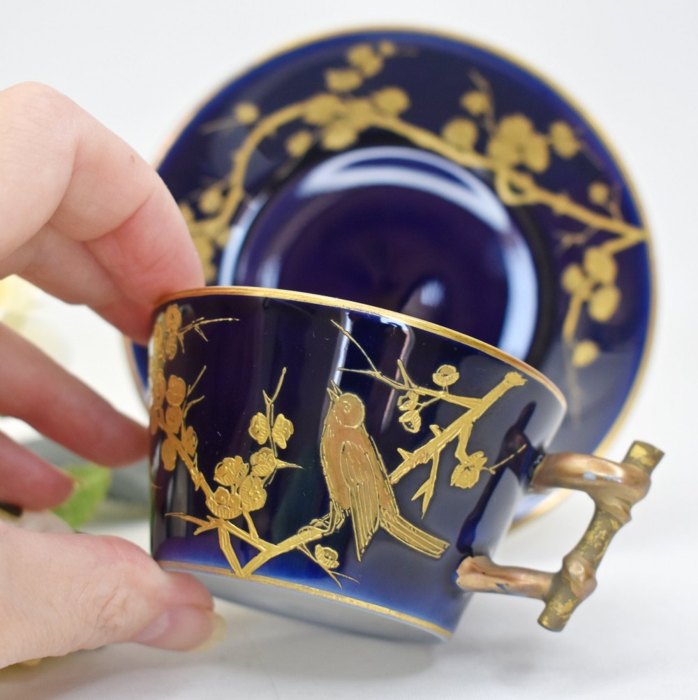 A dark cobalt blue porcelain coffee cup and matching saucer decorated with raised gilt floral branches, a perched bird, and a bamboo-style handle. View II