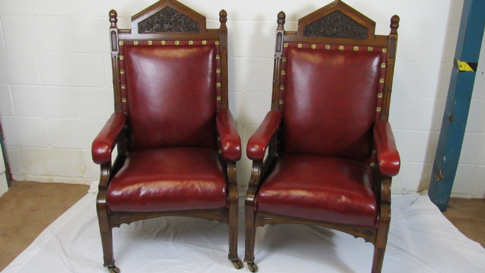 A pair of high-backed Gothic Revival armchairs crafted from carved dark wood and upholstered in deep red leather with brass stud detail.