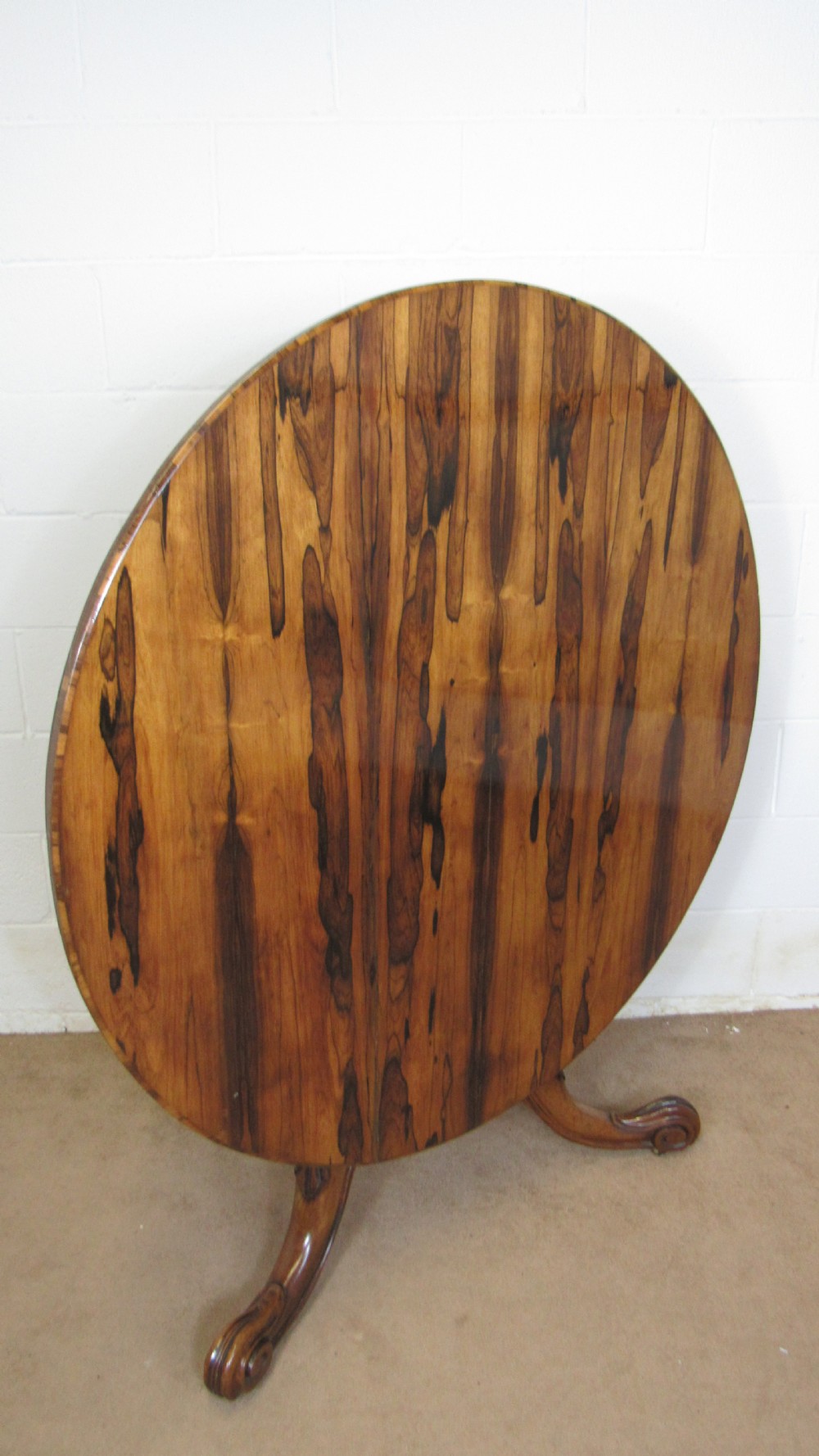 A circular antique rosewood tilt-top table shown in a vertical tilted position, displaying figured timber grain and a carved tripod base.