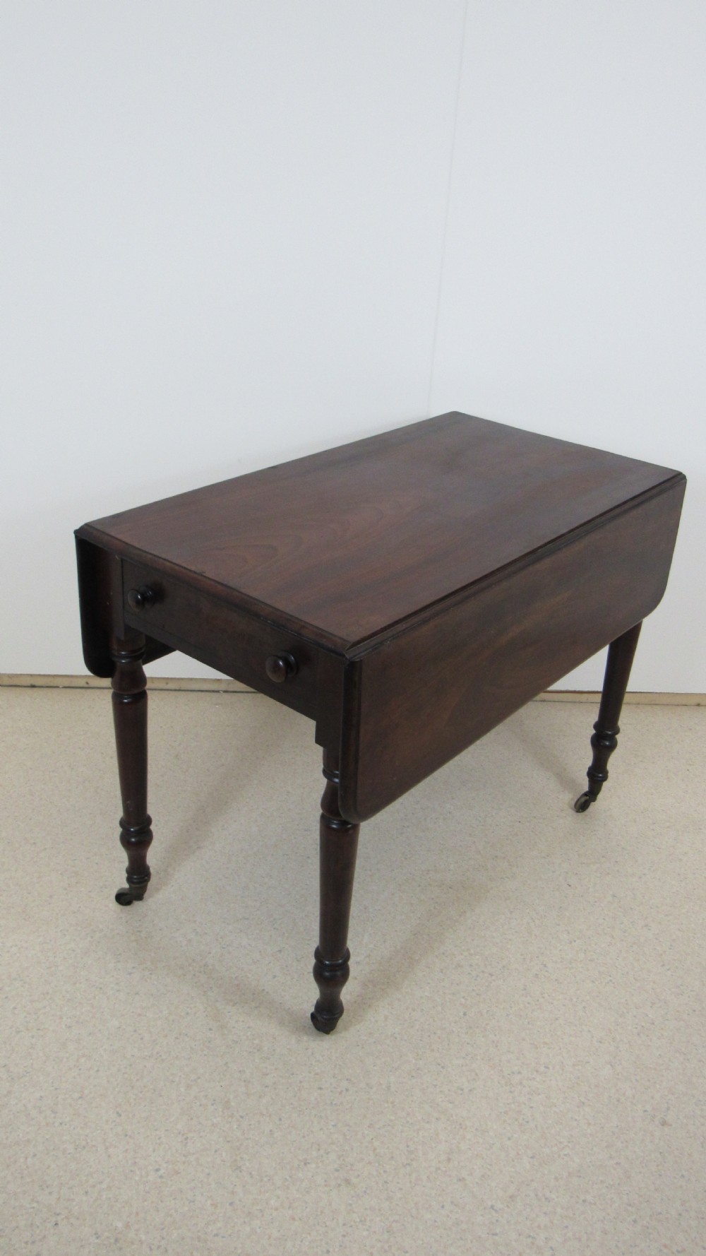 A dark mahogany Pembroke table with two drop leaves, turned legs on brass castors, and a single end drawer with wooden knobs.