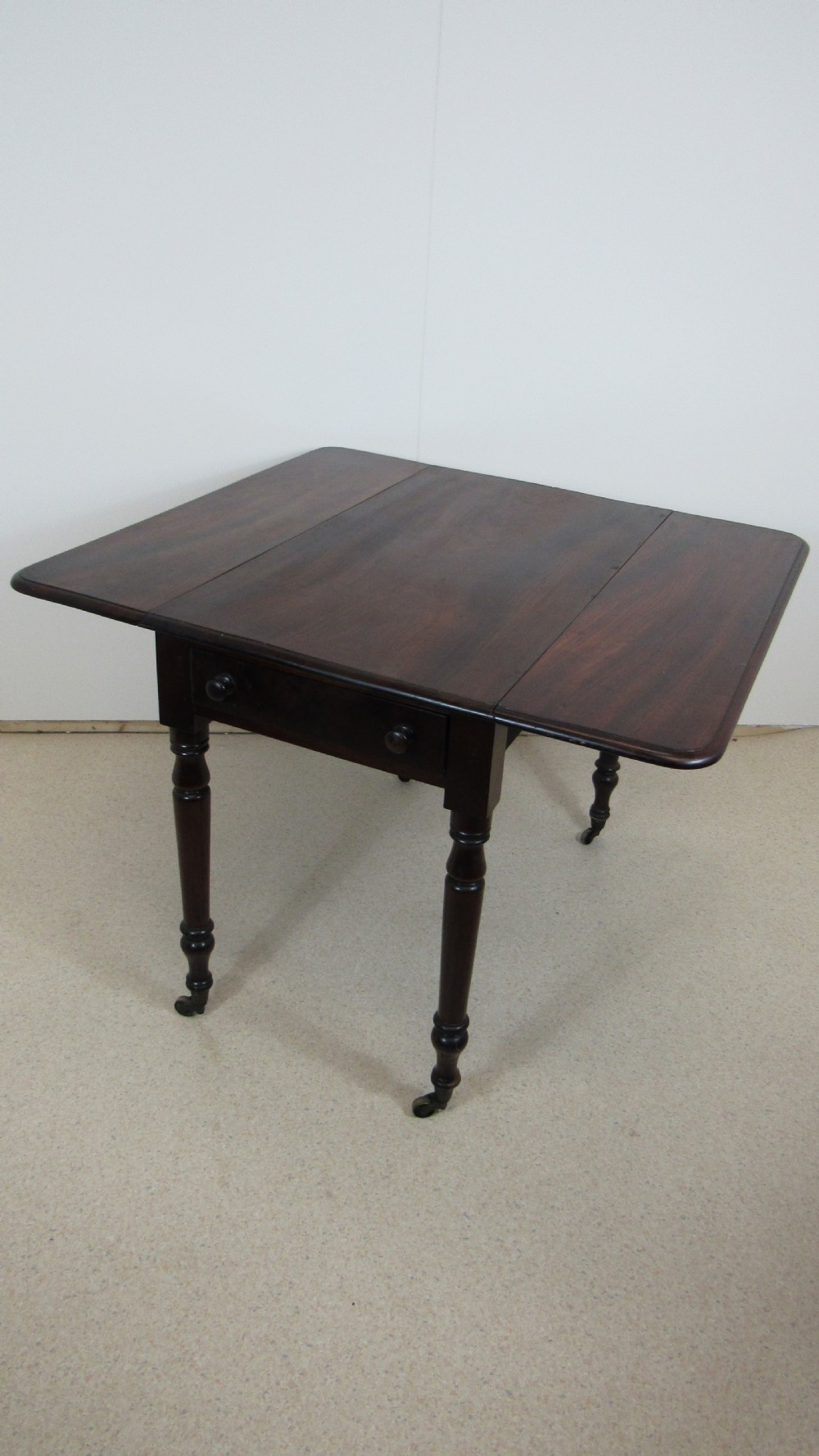 A dark mahogany Pembroke table with two drop leaves, turned legs on brass castors, and a single end drawer with wooden knobs. View VI