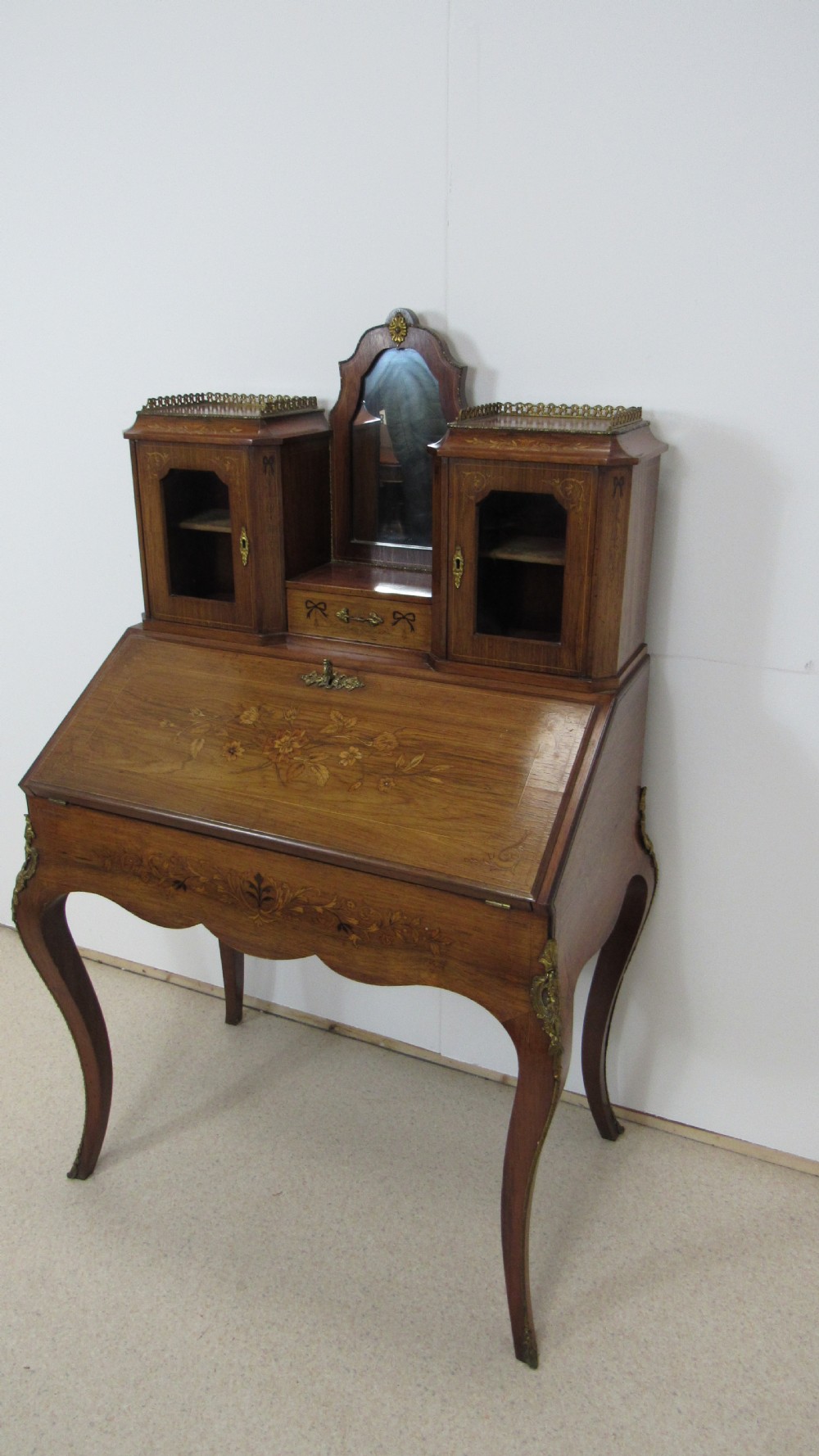 An antique French-style walnut writing bureau with floral marquetry, cabriole legs, brass mounts, and a upper cabinet section featuring small glazed doors and a central mirror.