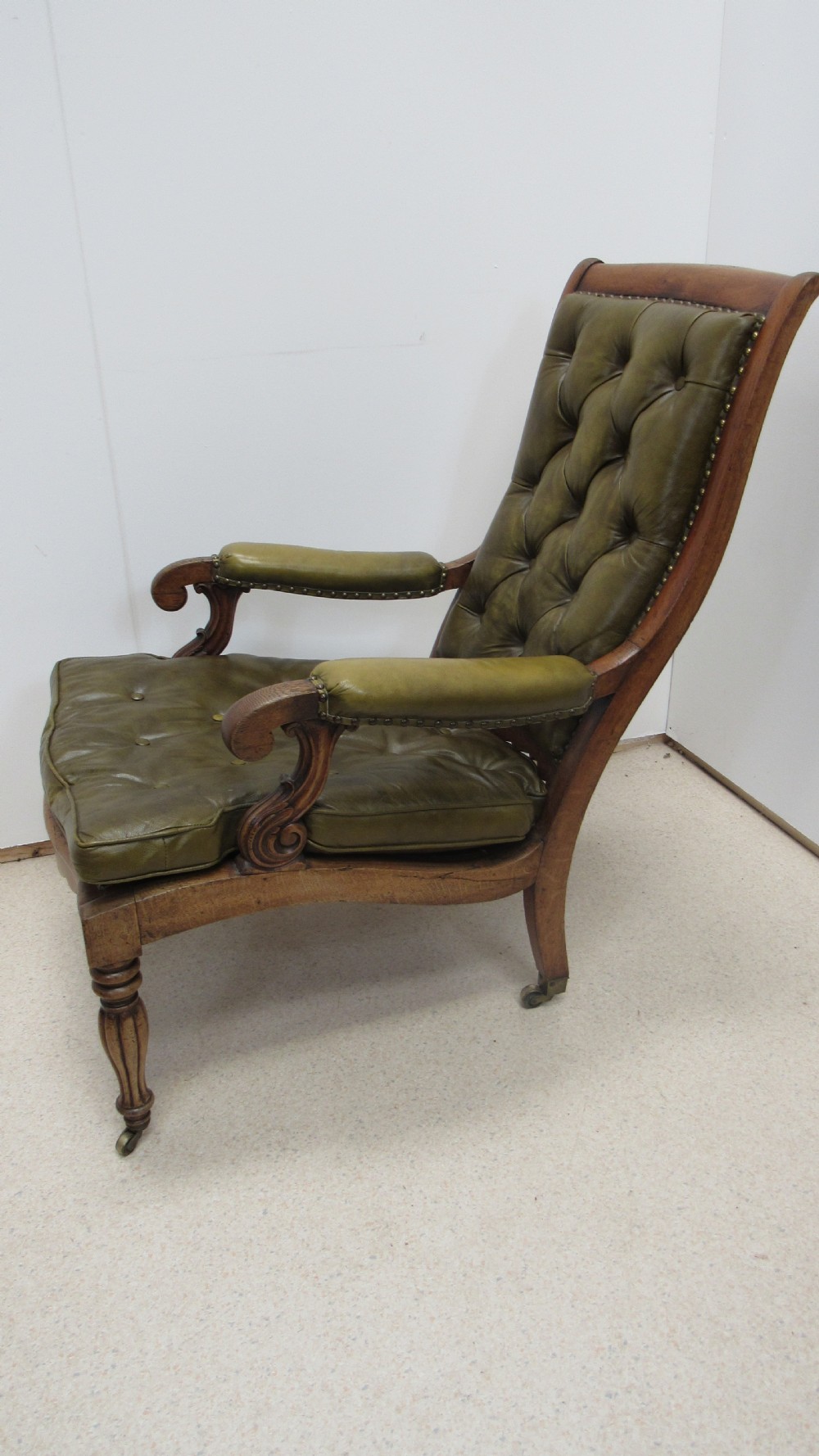 A William IV mahogany library chair upholstered in deep-buttoned olive green leather with padded scroll arms and turned front legs on brass castors. View X