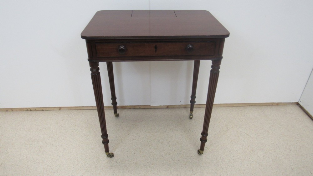 A Regency mahogany chamber table featuring reeded tapered legs on brass castors, a central drawer with wooden knobs, and a hinged lid top.