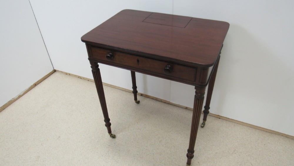 A Regency mahogany chamber table featuring reeded tapered legs on brass castors, a central drawer with wooden knobs, and a hinged lid top. View II