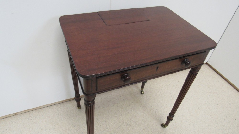 A Regency mahogany chamber table featuring reeded tapered legs on brass castors, a central drawer with wooden knobs, and a hinged lid top. View VI
