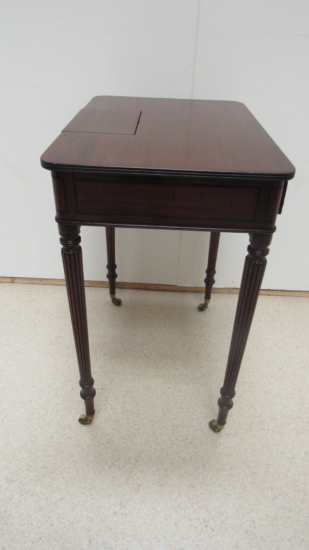 A Regency mahogany chamber table featuring reeded tapered legs on brass castors, a central drawer with wooden knobs, and a hinged lid top. View XIV