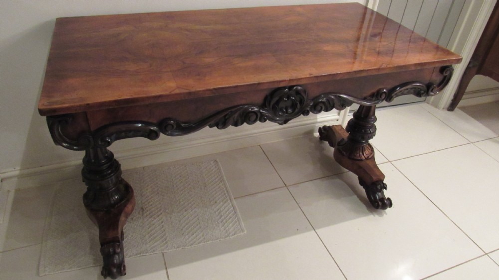 An antique Victorian walnut writing centre table featuring a figured top, deeply carved foliate apron, and carved turned end supports on a trestle base.