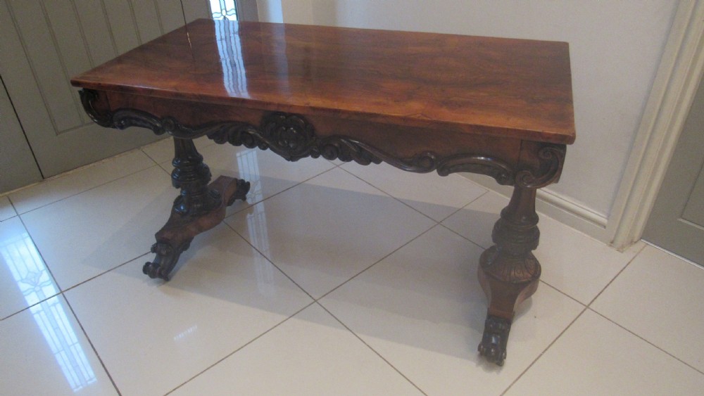 An antique Victorian walnut writing centre table featuring a figured top, deeply carved foliate apron, and carved turned end supports on a trestle base. View II