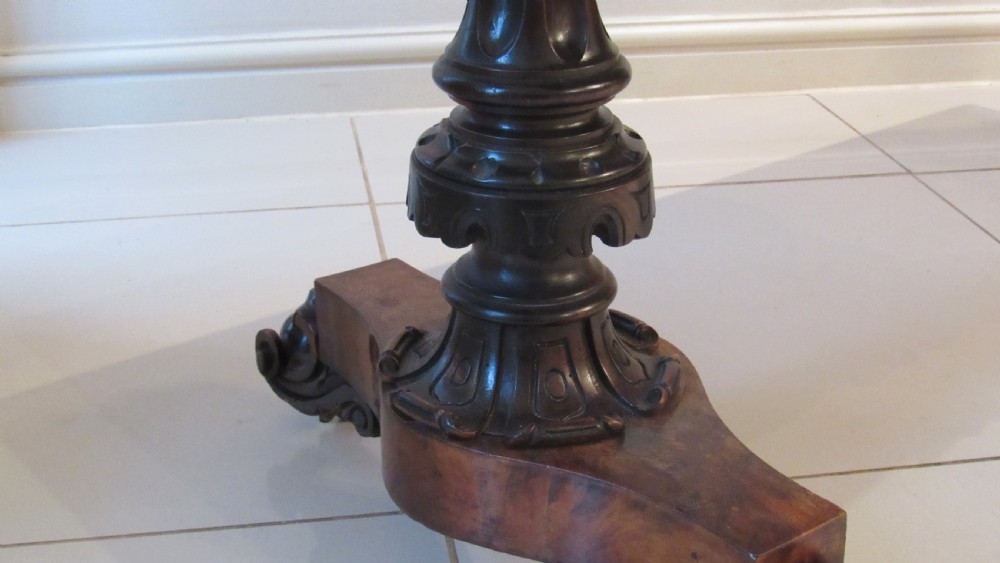 An antique Victorian walnut writing centre table featuring a figured top, deeply carved foliate apron, and carved turned end supports on a trestle base. View VI
