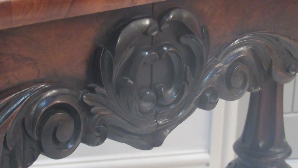 An antique Victorian walnut writing centre table featuring a figured top, deeply carved foliate apron, and carved turned end supports on a trestle base. View X
