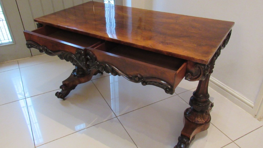 An antique Victorian walnut writing centre table featuring a figured top, deeply carved foliate apron, and carved turned end supports on a trestle base. View XI