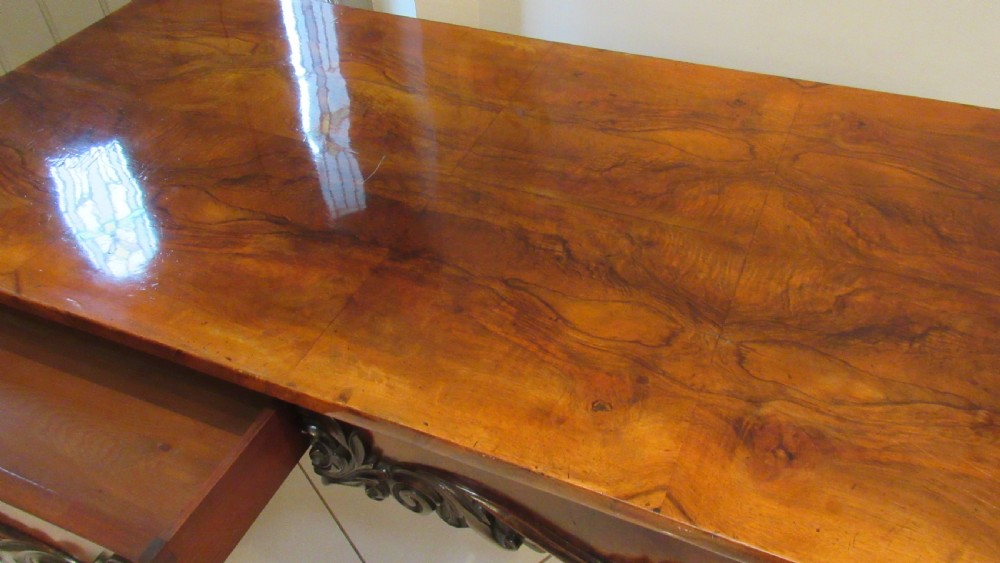 An antique Victorian walnut writing centre table featuring a figured top, deeply carved foliate apron, and carved turned end supports on a trestle base. View XII