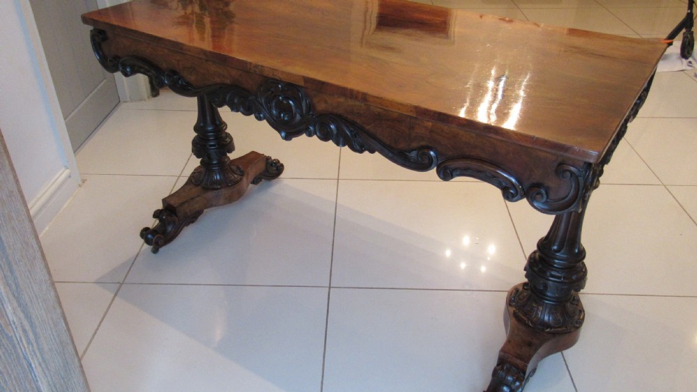 An antique Victorian walnut writing centre table featuring a figured top, deeply carved foliate apron, and carved turned end supports on a trestle base. View XIV