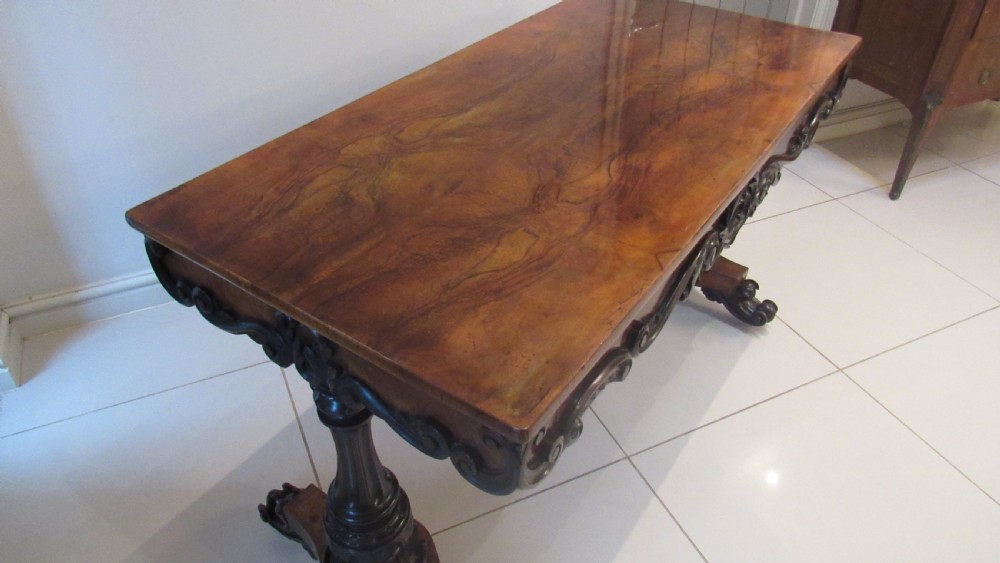 An antique Victorian walnut writing centre table featuring a figured top, deeply carved foliate apron, and carved turned end supports on a trestle base. View XV