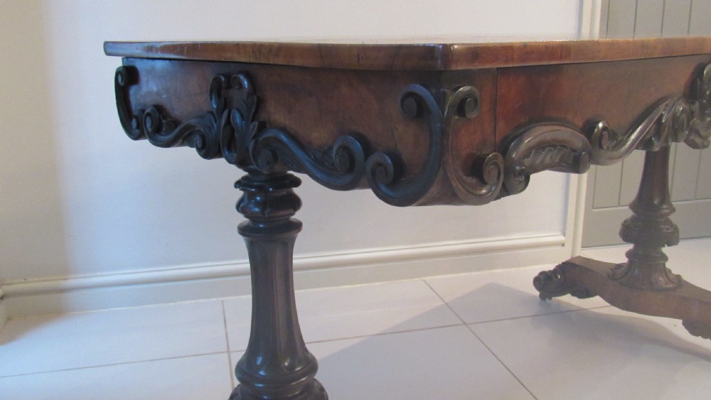 An antique Victorian walnut writing centre table featuring a figured top, deeply carved foliate apron, and carved turned end supports on a trestle base. View XVI