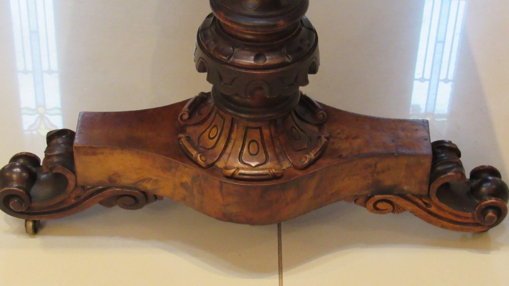 An antique Victorian walnut writing centre table featuring a figured top, deeply carved foliate apron, and carved turned end supports on a trestle base. View XIX