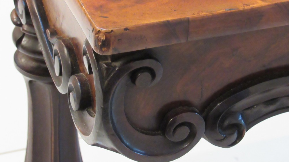 An antique Victorian walnut writing centre table featuring a figured top, deeply carved foliate apron, and carved turned end supports on a trestle base. View XX