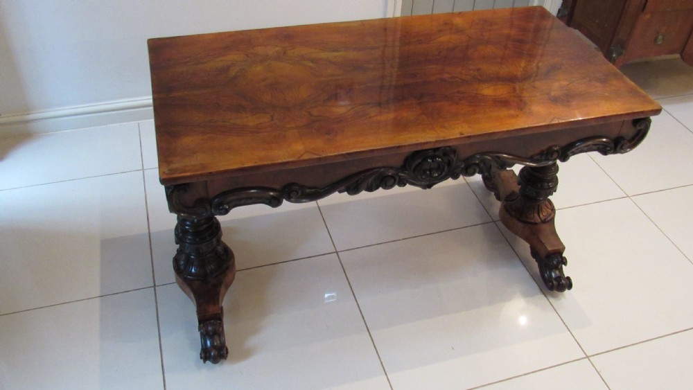 An antique Victorian walnut writing centre table featuring a figured top, deeply carved foliate apron, and carved turned end supports on a trestle base. View XXII