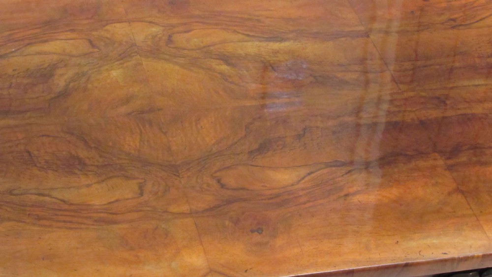 An antique Victorian walnut writing centre table featuring a figured top, deeply carved foliate apron, and carved turned end supports on a trestle base. View XXIV