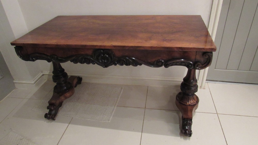 An antique Victorian walnut writing centre table featuring a figured top, deeply carved foliate apron, and carved turned end supports on a trestle base. View XXV