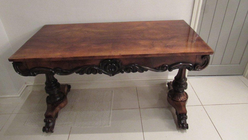 An antique Victorian walnut writing centre table featuring a figured top, deeply carved foliate apron, and carved turned end supports on a trestle base. View XXVI