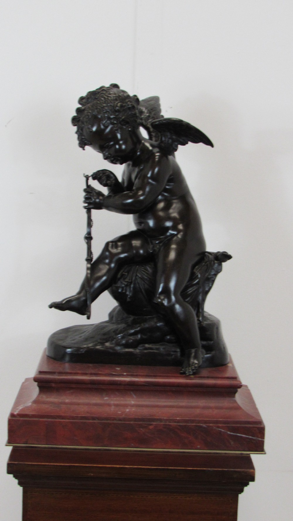 A dark patinated bronze figure of a winged cherub seated on a rock while holding a bow, set upon a rectangular reddish marble plinth.