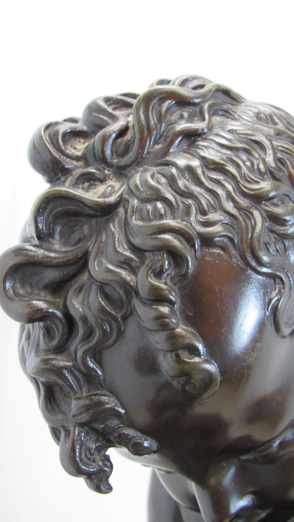 A dark patinated bronze figure of a winged cherub seated on a rock while holding a bow, set upon a rectangular reddish marble plinth. View V