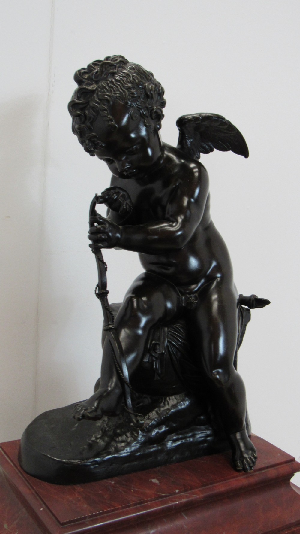 A dark patinated bronze figure of a winged cherub seated on a rock while holding a bow, set upon a rectangular reddish marble plinth. View VII