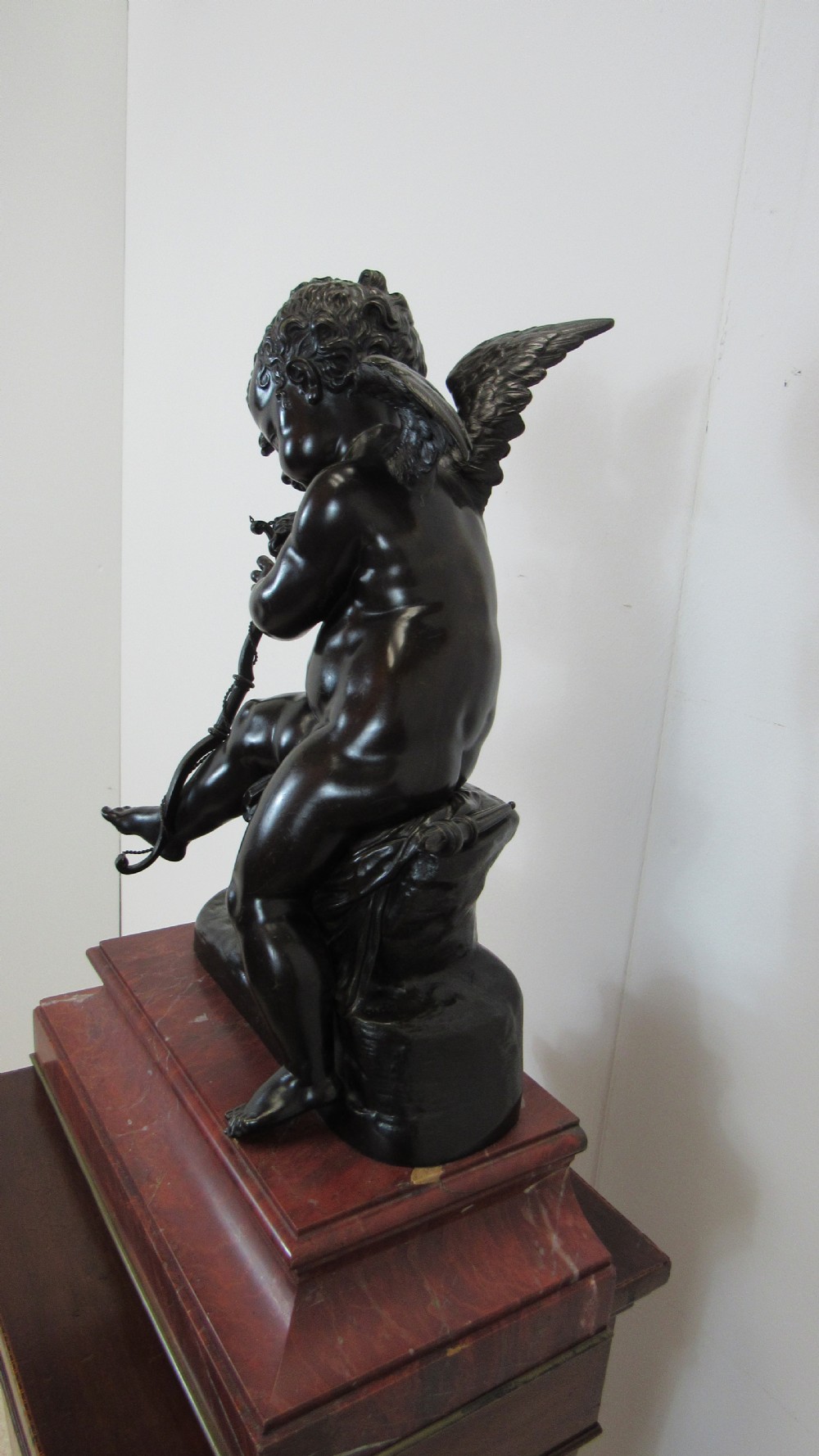 A dark patinated bronze figure of a winged cherub seated on a rock while holding a bow, set upon a rectangular reddish marble plinth. View IX