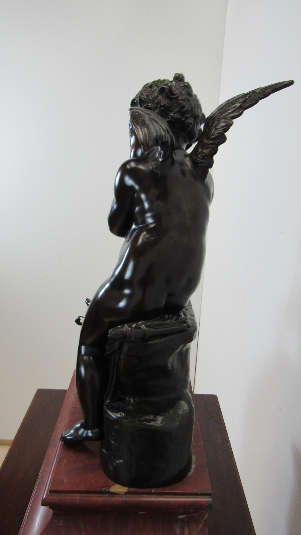 A dark patinated bronze figure of a winged cherub seated on a rock while holding a bow, set upon a rectangular reddish marble plinth. View X