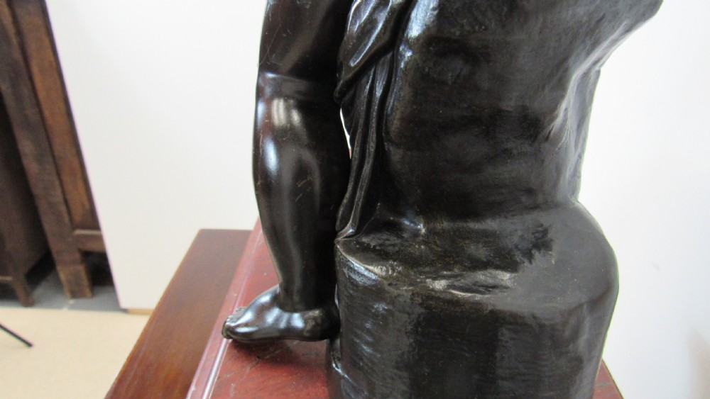A dark patinated bronze figure of a winged cherub seated on a rock while holding a bow, set upon a rectangular reddish marble plinth. View XII