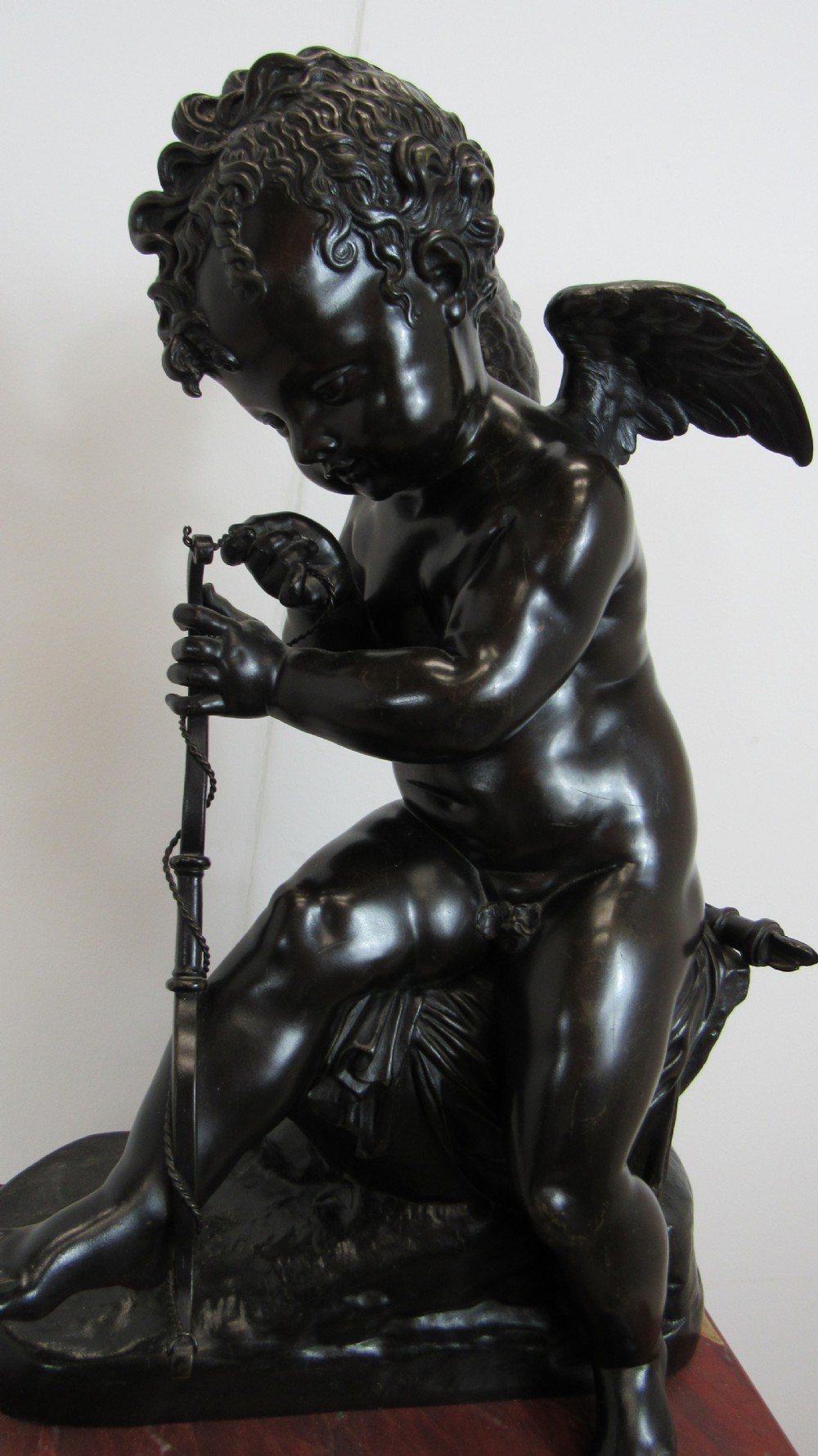 A dark patinated bronze figure of a winged cherub seated on a rock while holding a bow, set upon a rectangular reddish marble plinth. View XIII