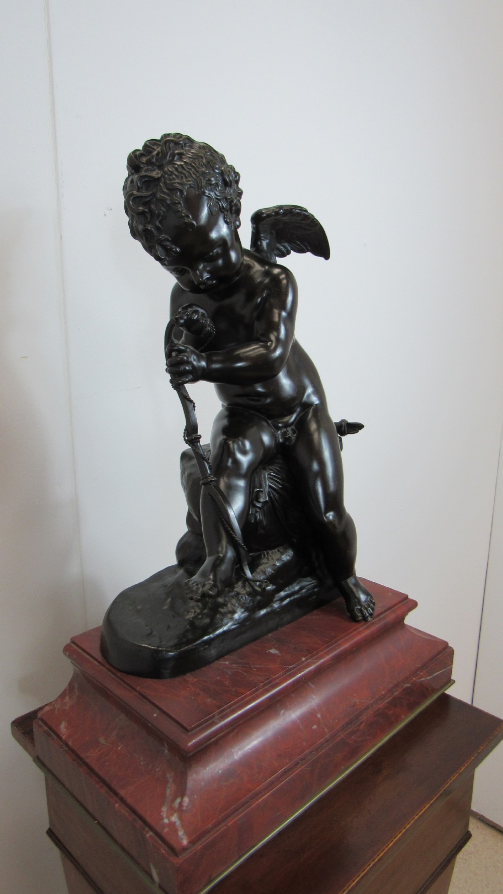 A dark patinated bronze figure of a winged cherub seated on a rock while holding a bow, set upon a rectangular reddish marble plinth. View XIV