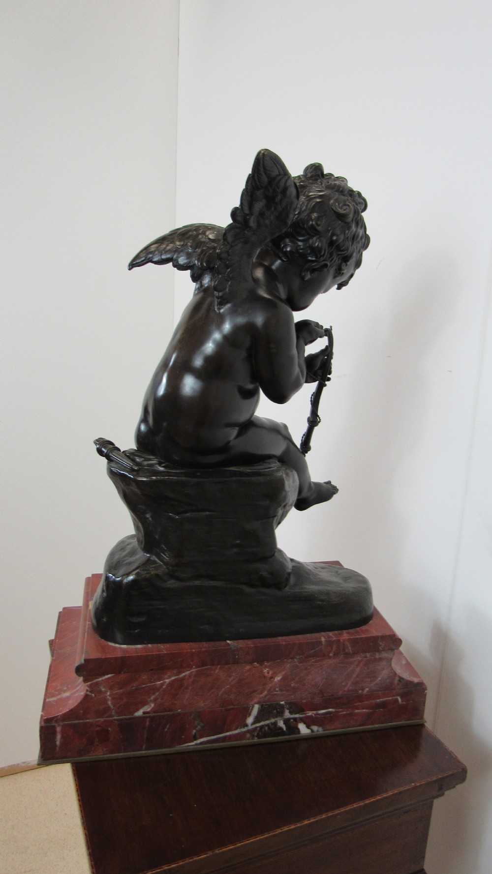 A dark patinated bronze figure of a winged cherub seated on a rock while holding a bow, set upon a rectangular reddish marble plinth. View XV