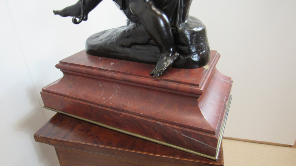 A dark patinated bronze figure of a winged cherub seated on a rock while holding a bow, set upon a rectangular reddish marble plinth. View XVI