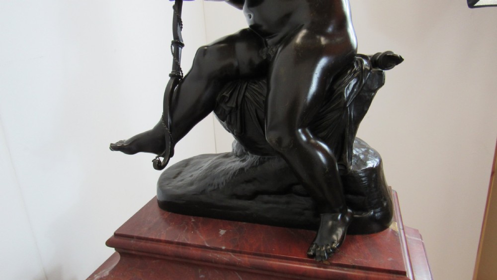A dark patinated bronze figure of a winged cherub seated on a rock while holding a bow, set upon a rectangular reddish marble plinth. View XVII