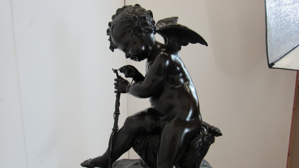 A dark patinated bronze figure of a winged cherub seated on a rock while holding a bow, set upon a rectangular reddish marble plinth. View XVIII
