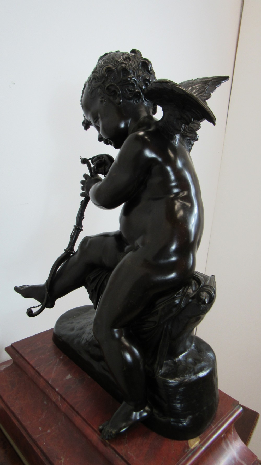 A dark patinated bronze figure of a winged cherub seated on a rock while holding a bow, set upon a rectangular reddish marble plinth. View XIX