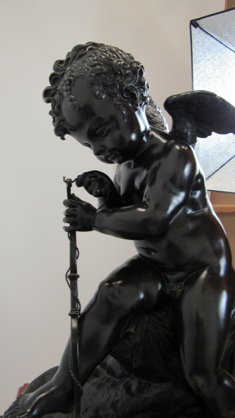 A dark patinated bronze figure of a winged cherub seated on a rock while holding a bow, set upon a rectangular reddish marble plinth. View XX