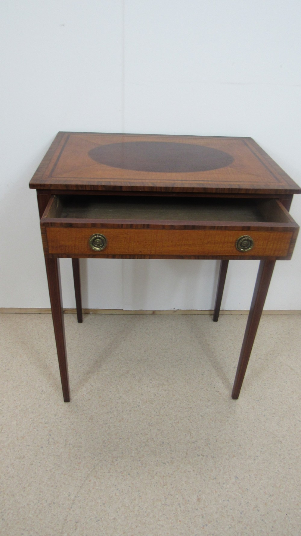 A 19th-century wooden side table featuring square tapered legs, a single drawer with brass handles, and a decorative oval inlaid top surface. View X